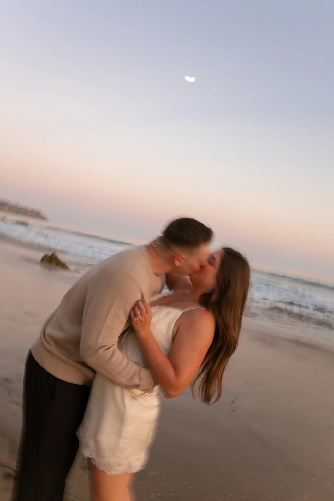 A couple kissing on the beach with the woman lifting one leg and wrapping her arms around her partner during golden hour.