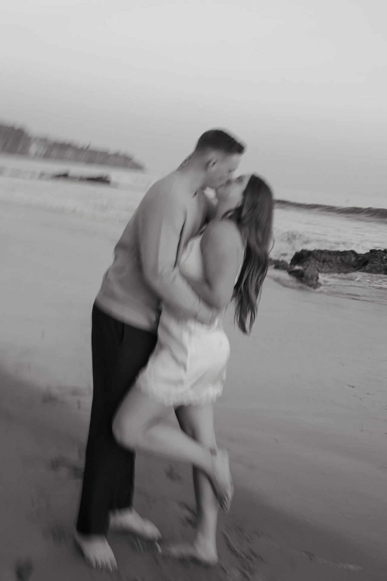 Beach Engagement Photo Ideas of a blurred motion shot of a couple kissing on the beach, the woman lifting one leg as waves and sand create a dreamy effect.