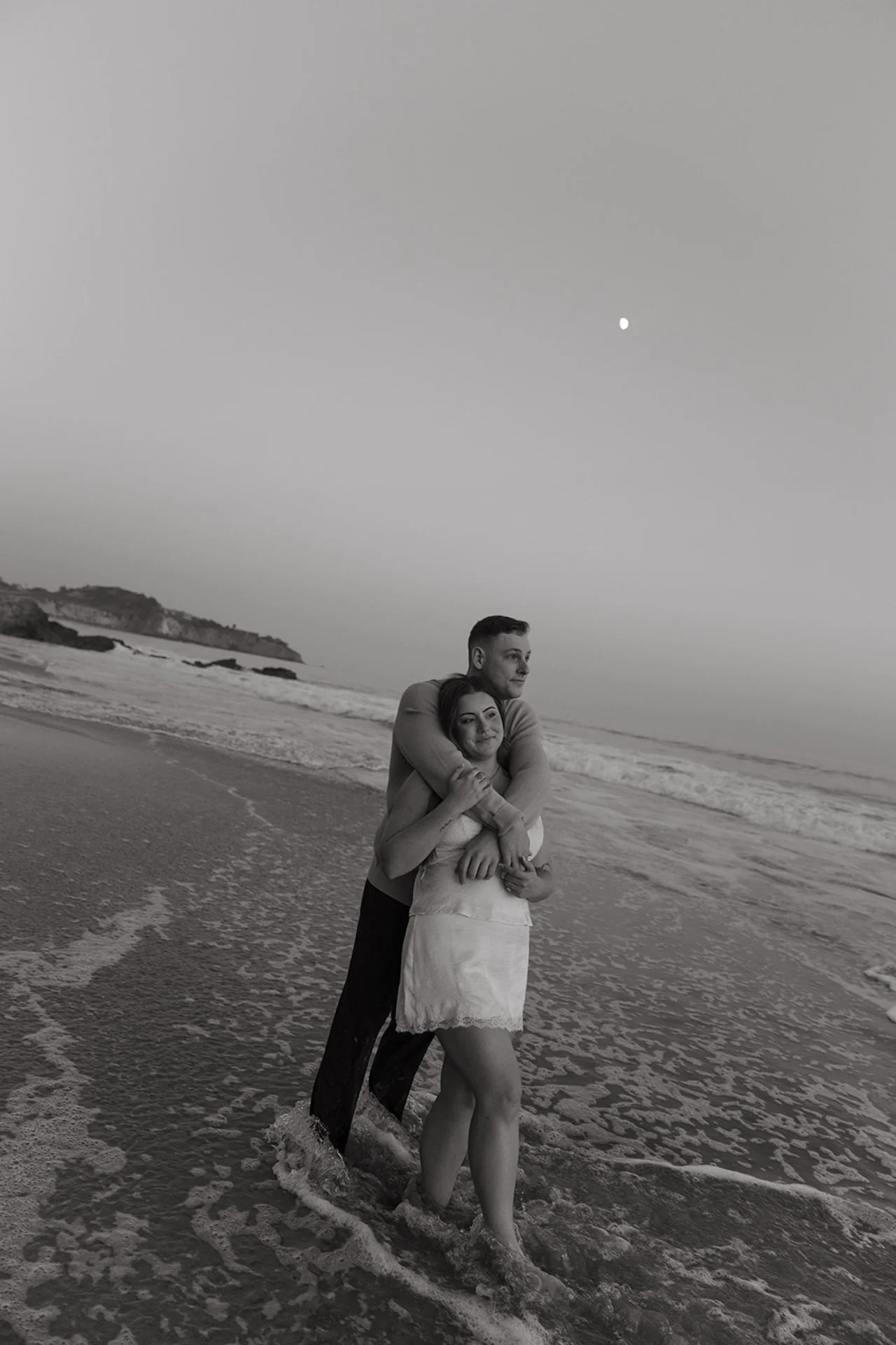 A couple standing in the shallow water in black and white, the man hugging the woman from behind while waves surround their feet and the moon hangs in the sky.