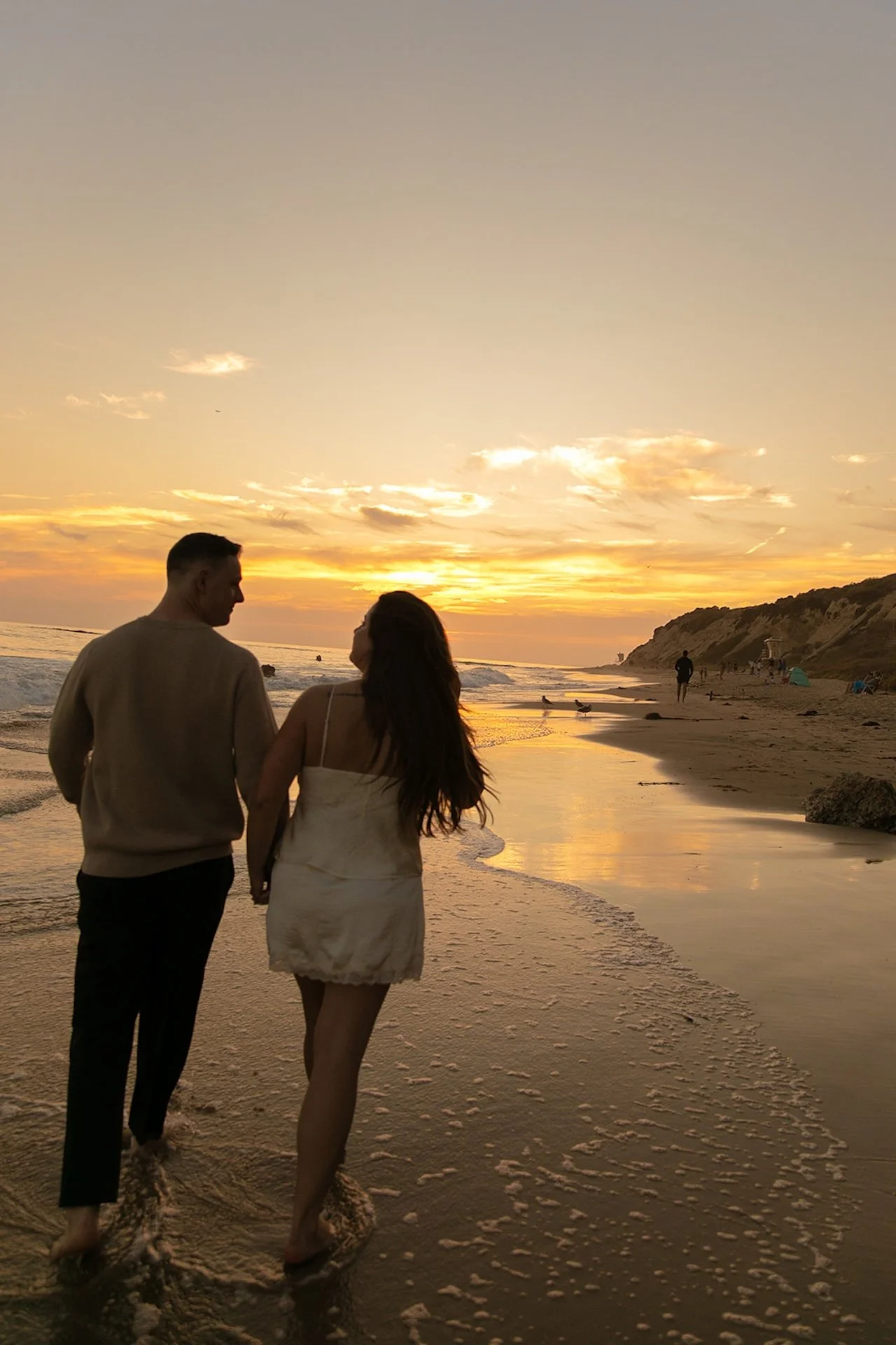 A wide shot of a couple walking along the shoreline at sunset, holding hands with reflections in the wet sand and cliffs in the distance.