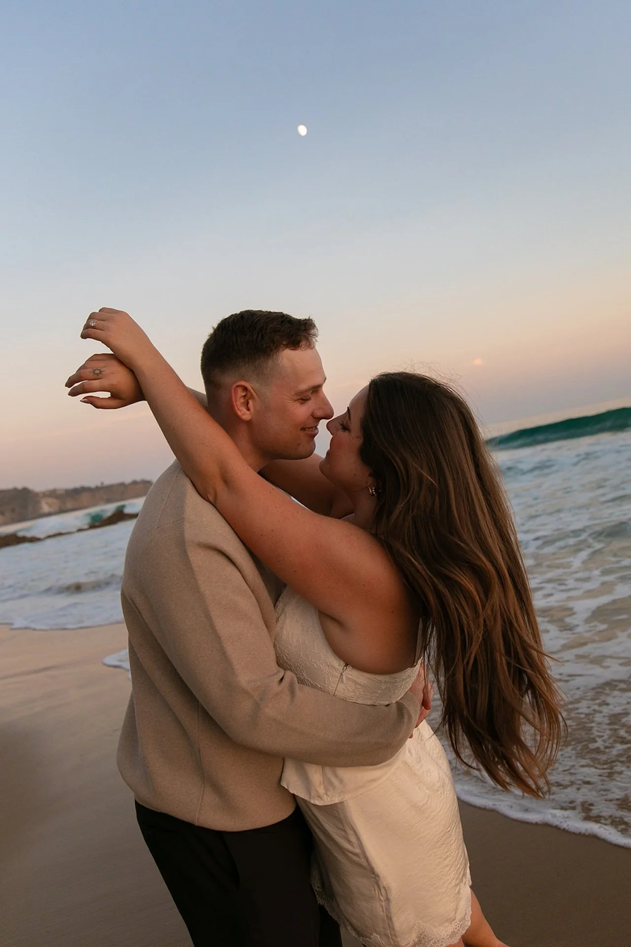 A close-up of a couple hugging on the beach at sunset, the woman wrapping her arms around the man’s neck with the ocean and moon in the background.