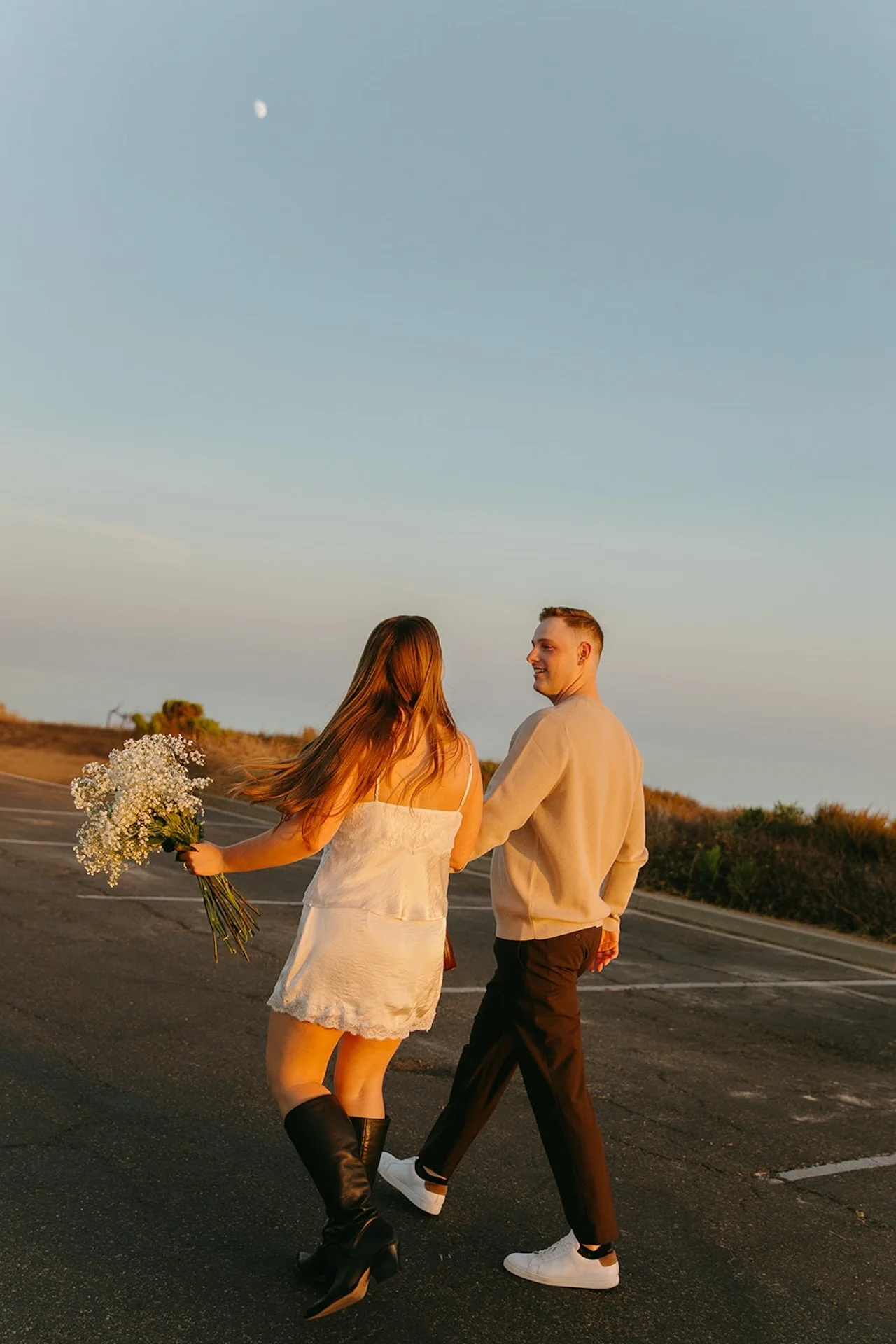 A couple walking hand in hand on a coastal road at golden hour, the woman holding a bouquet of baby’s breath while both look back toward the camera.