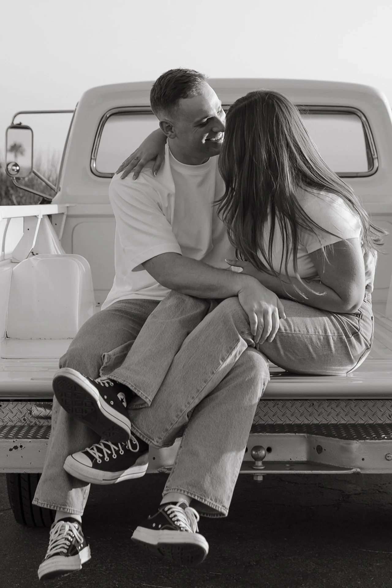 Beach Engagement Photo Ideas of a couple sitting on the tailgate of a vintage white truck, leaning in close and smiling at each other in casual white shirts and denim at golden hour.