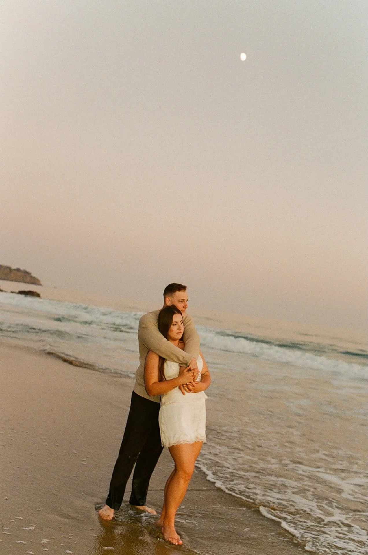 A couple standing in the ocean at sunset, the man hugging the woman from behind while waves roll in and the moon appears in the pastel sky.