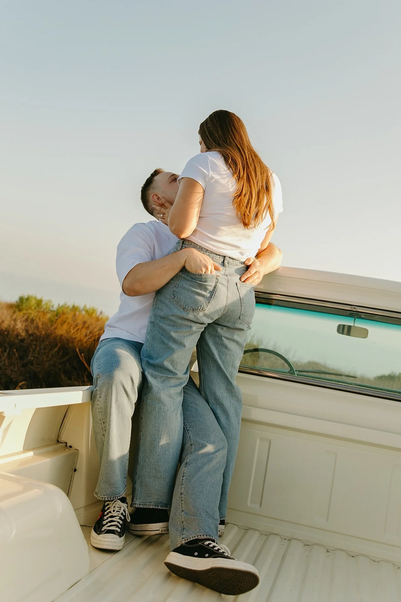A wide shot of a couple standing in the bed of a classic truck, embracing and kissing with soft golden-hour light and an open sky backdrop.