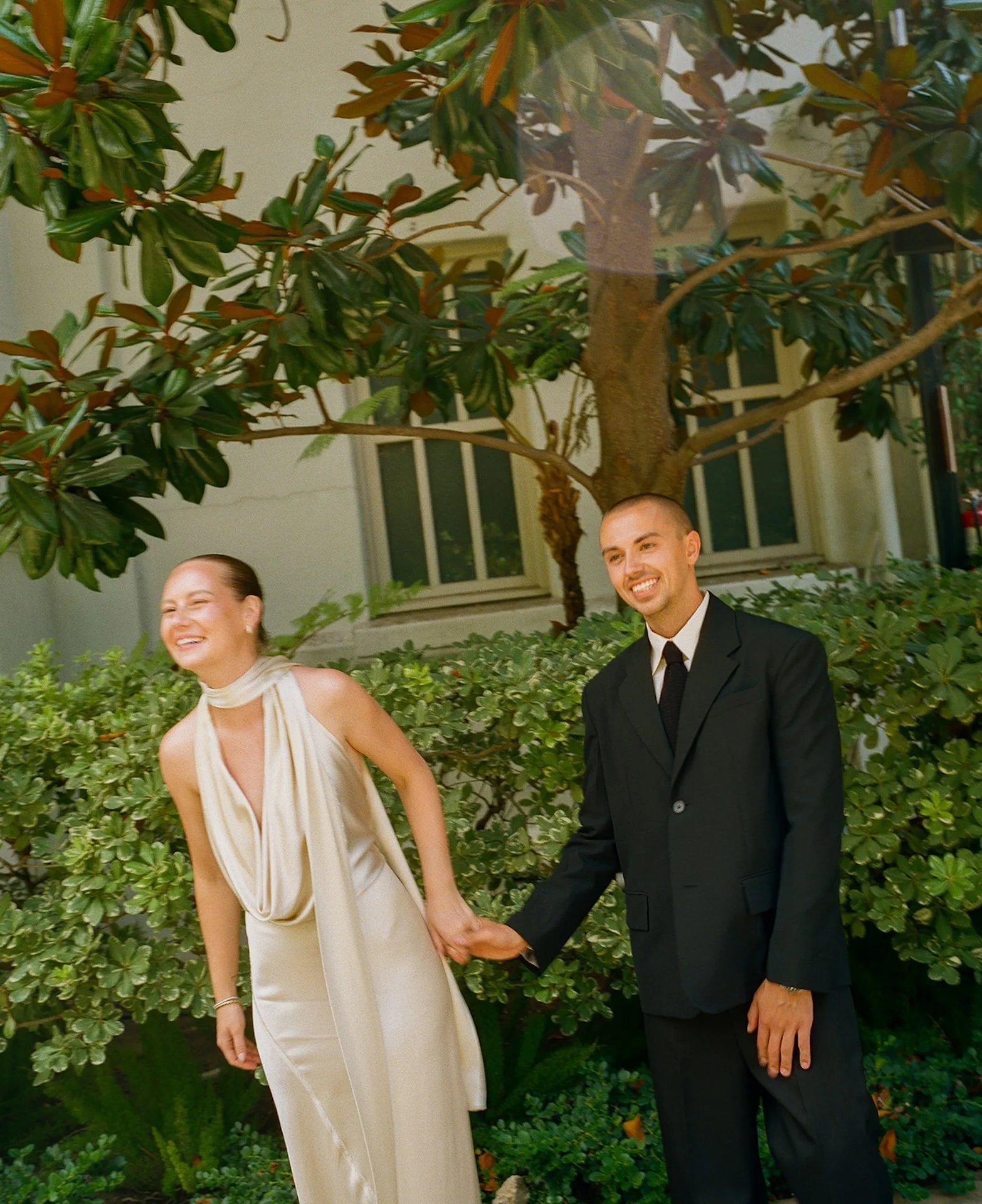 The couple holding hands and laughing together outdoors, walking through a garden setting after their intimate wedding ceremony.
