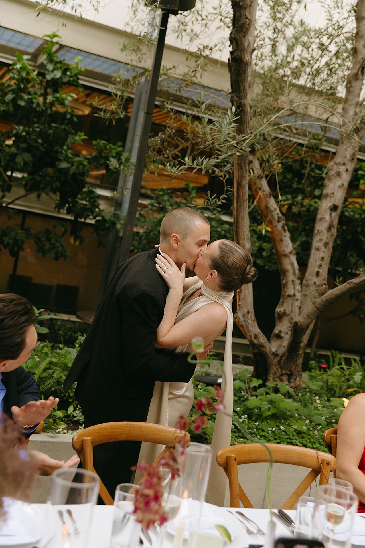 The couple sharing their first kiss during the ceremony in a garden courtyard with guests applauding from their seats.