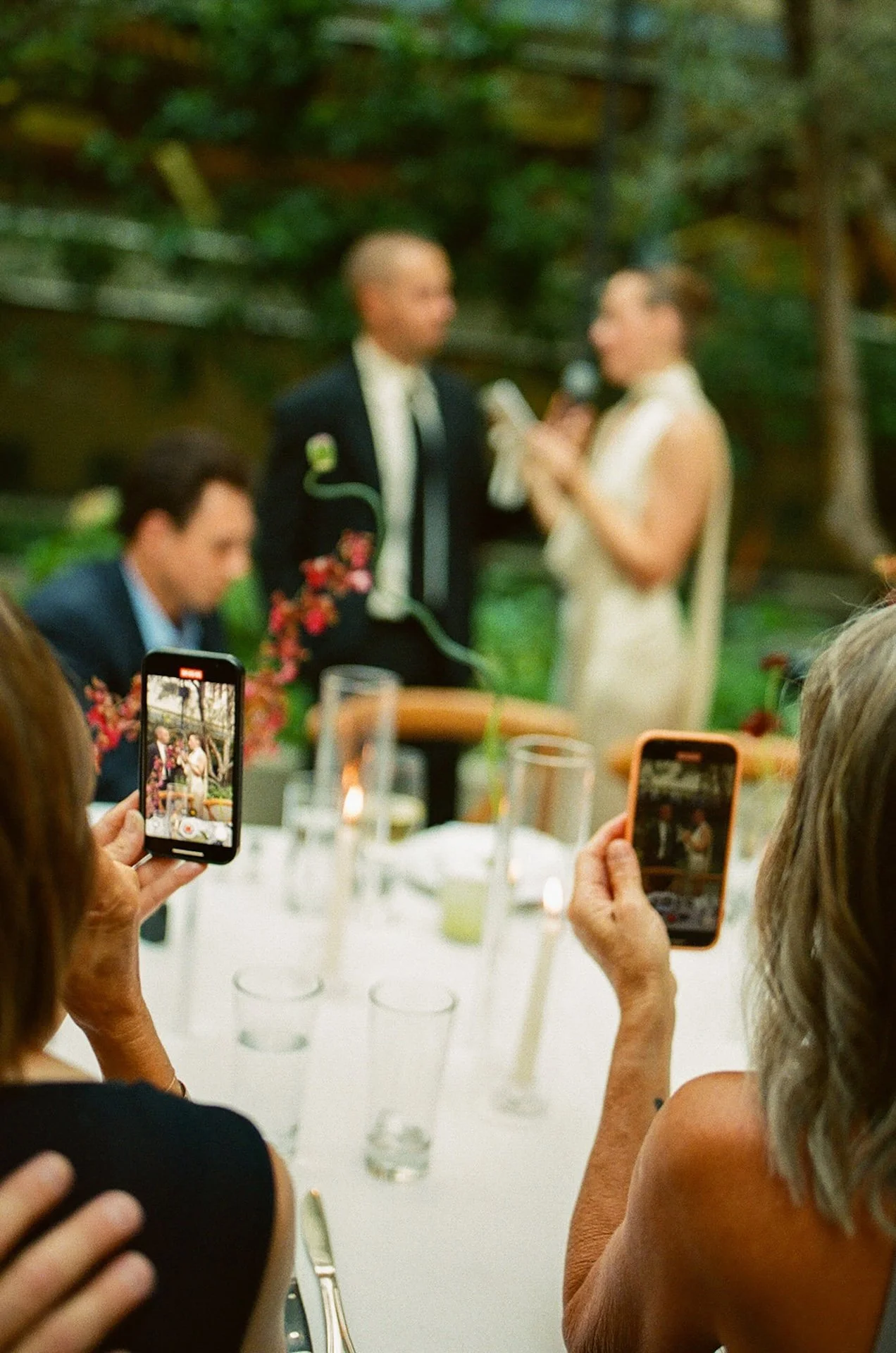 Guests recording the couple during their vows on their phones, highlighting Intimate Wedding Ideas with a documentary-style perspective at an outdoor dinner reception.