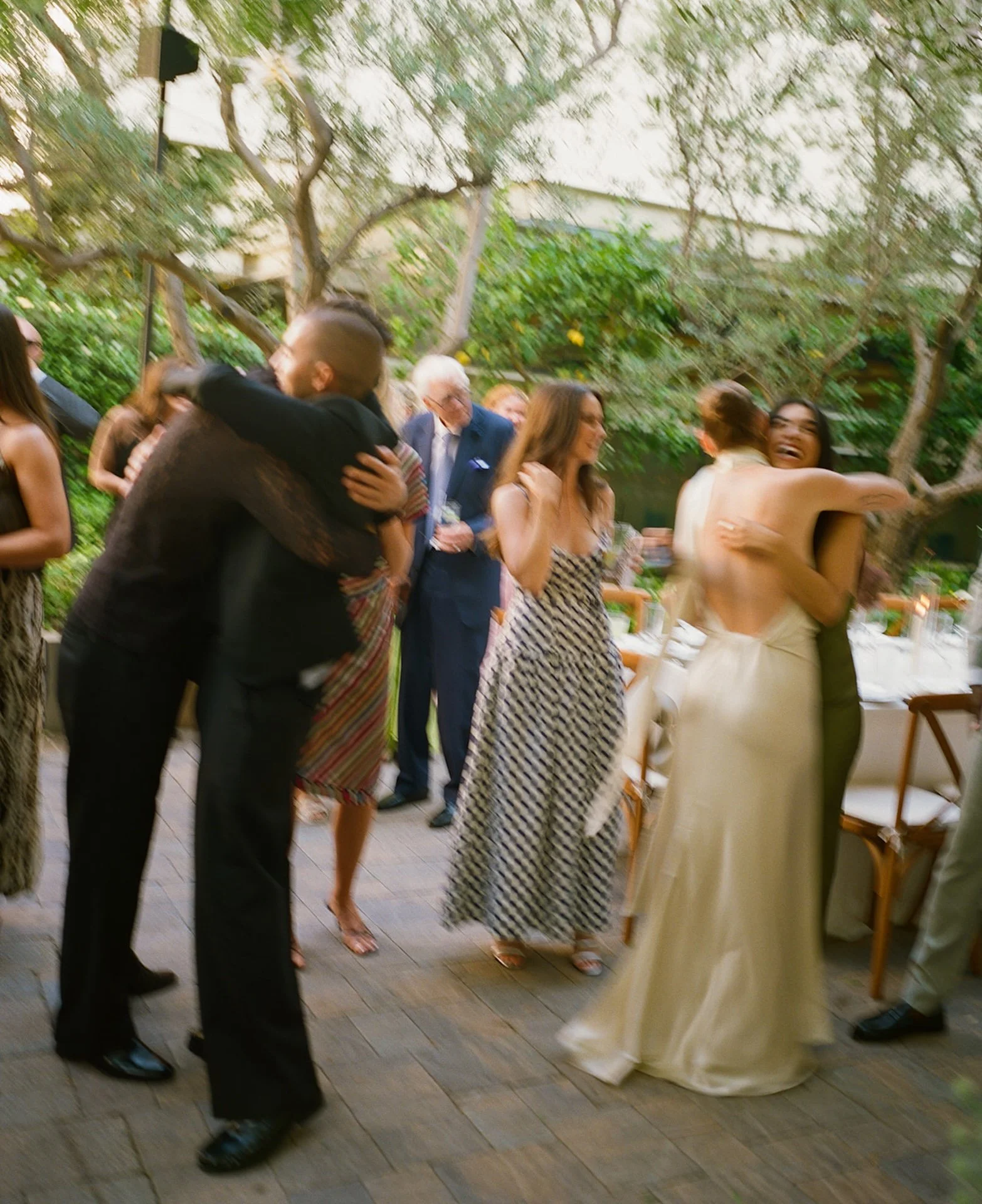 The couple hugging friends and guests during the reception, with blurred motion and greenery creating a lively, documentary-style wedding scene.
