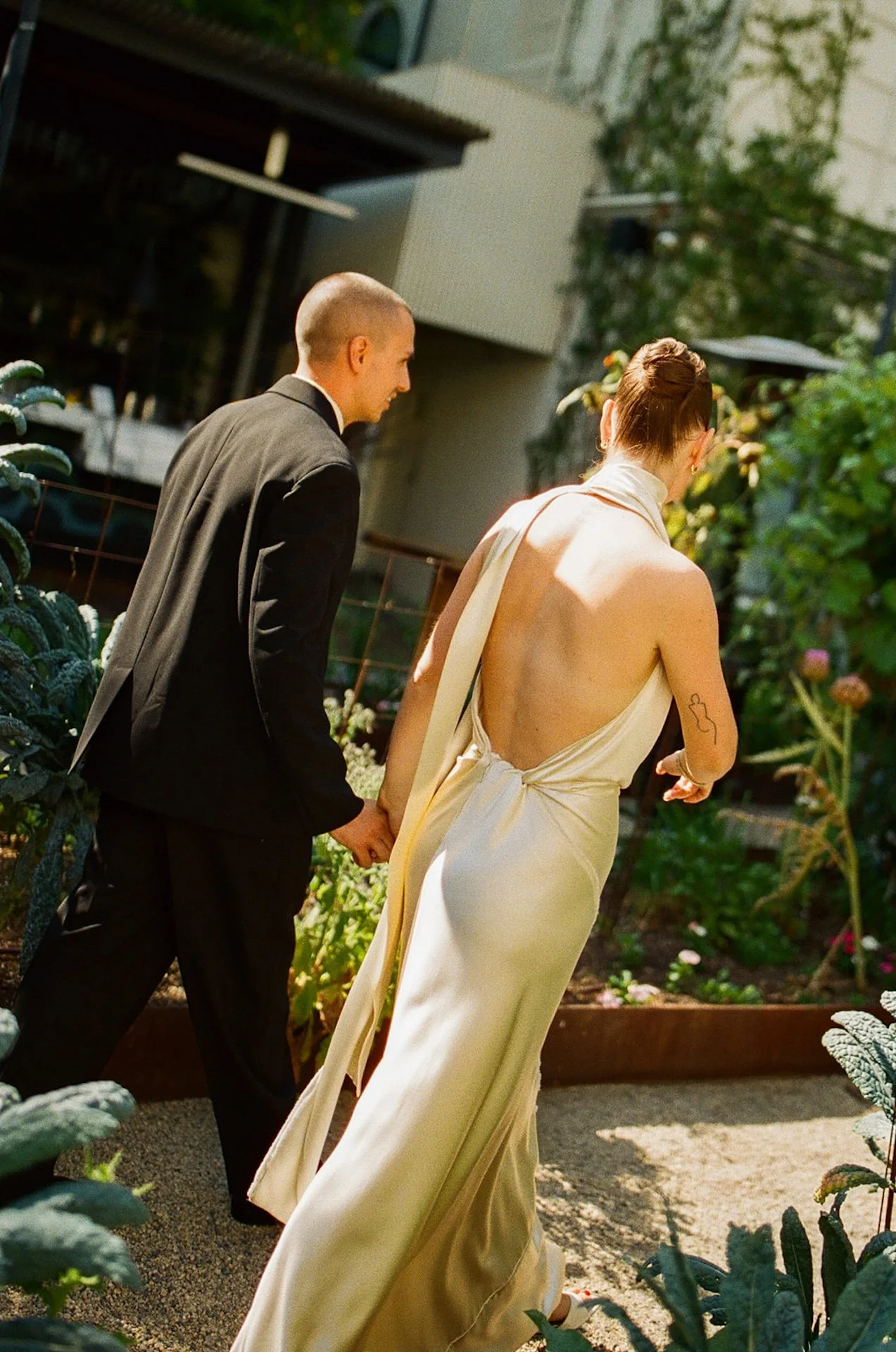 The bride and groom walking hand in hand through a garden path, with the bride wearing a backless satin gown and the groom in a black suit.