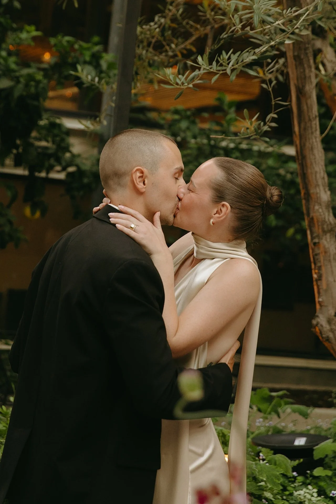 The couple sharing a kiss during their ceremony in a garden setting with olive trees and soft natural light.
