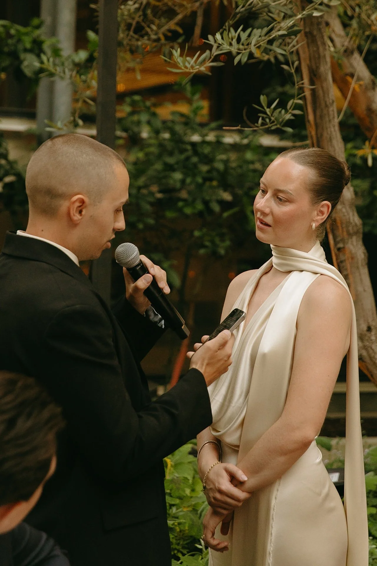 The groom reading vows into a microphone while the bride listens, both standing in a lush garden courtyard surrounded by greenery.