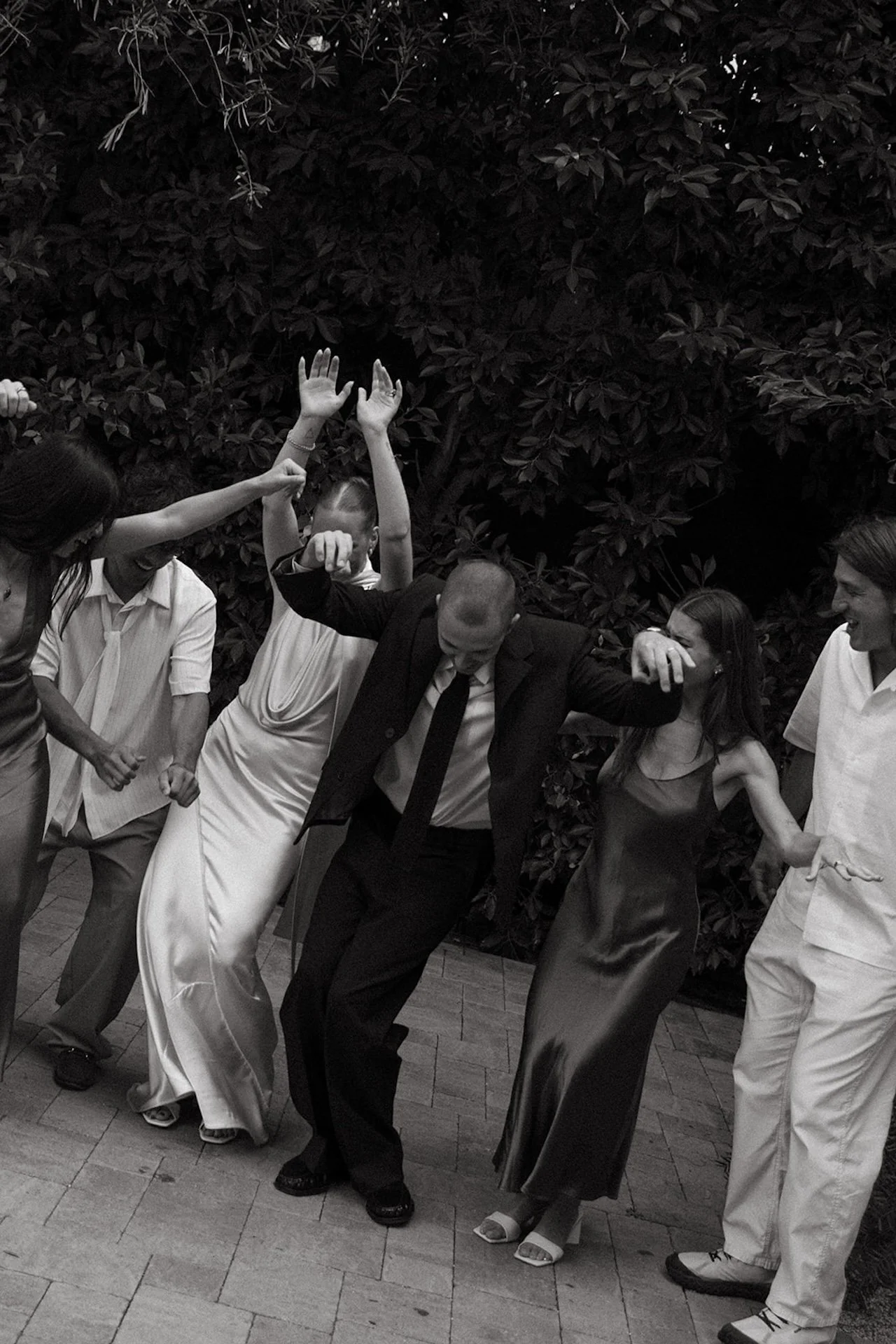 A group of guests dancing together outdoors during the wedding reception, with arms raised and candid movement in a garden courtyard setting.