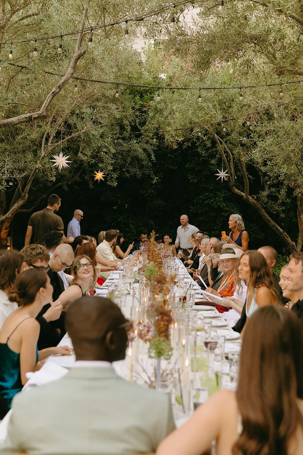 A long outdoor dinner table filled with guests under olive trees and string lights, showcasing Intimate Wedding Ideas for an alfresco wedding reception with candles and floral centerpieces.