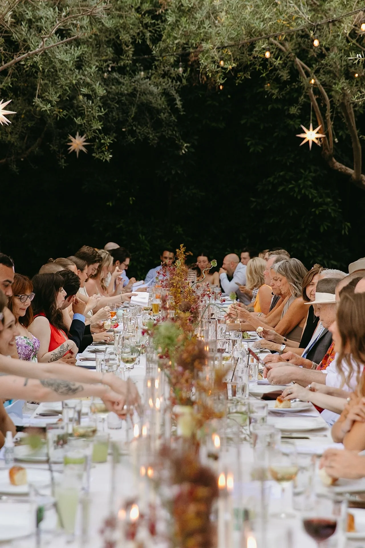 A long banquet table filled with guests dining outdoors under string lights and trees, highlighting Intimate Wedding Ideas for an elegant garden dinner reception.