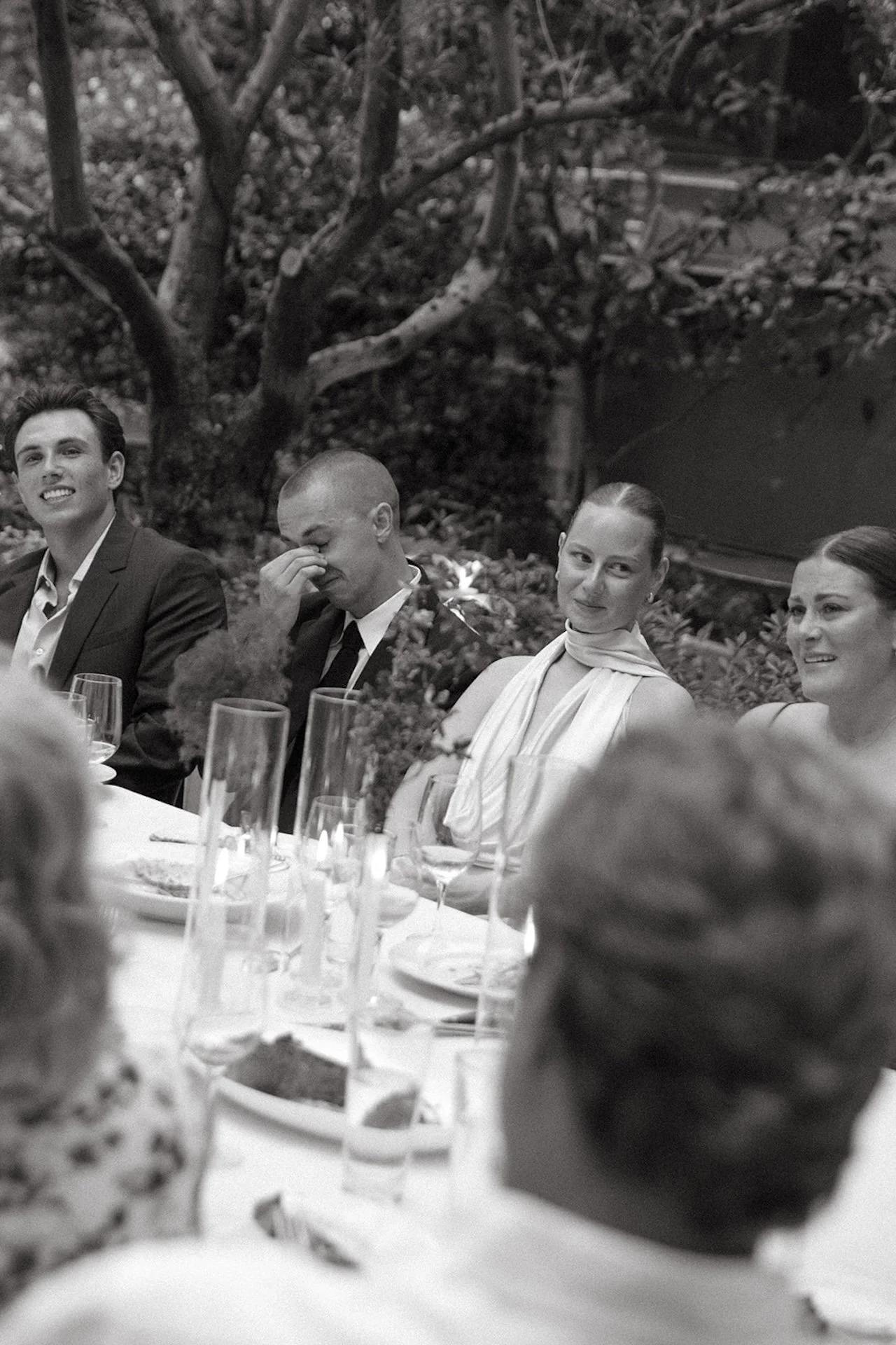 A black-and-white photo of the couple seated at the dinner table during speeches, smiling and reacting to toasts surrounded by guests and candlelight.
