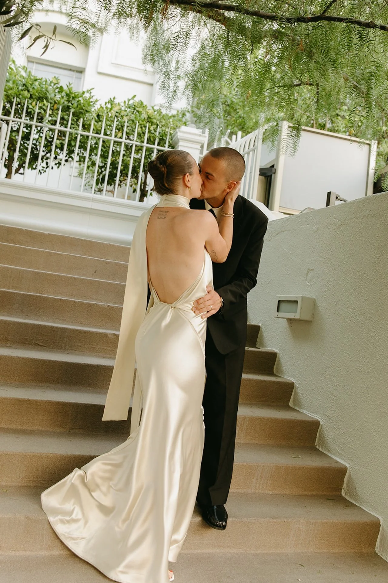 The bride and groom kissing on outdoor steps, with the bride in a backless satin gown and the groom in a black suit, surrounded by greenery and soft light.