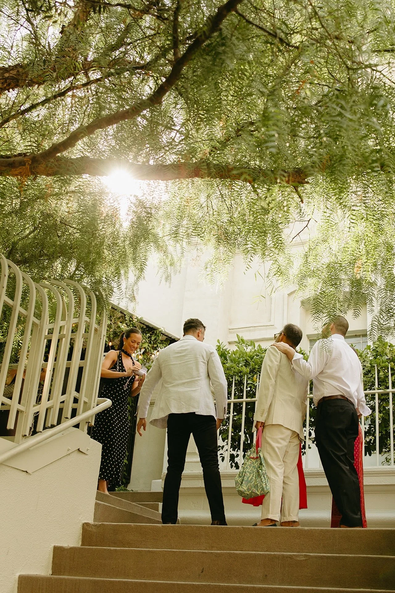 A wide shot of guests walking up steps into the venue under leafy trees and warm sunlight, showcasing Intimate Wedding Ideas for a garden-style city wedding setting.