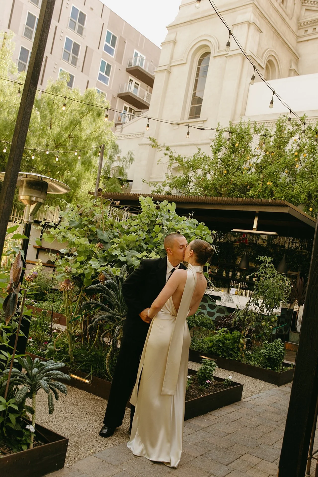 The couple kissing in a lush garden courtyard surrounded by raised garden beds, greenery, and string lights with an urban backdrop.