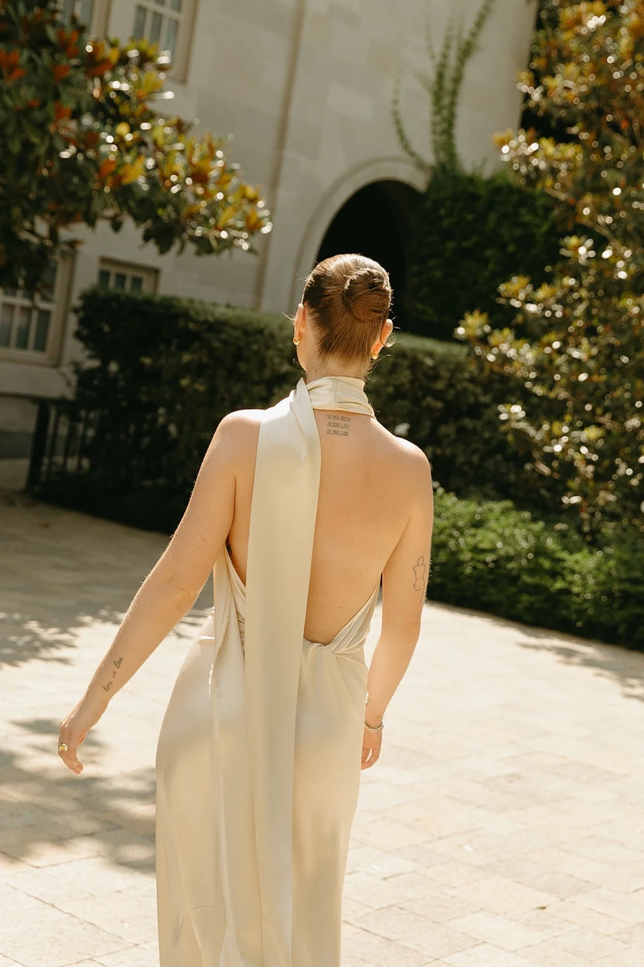 The bride walking through a sunlit courtyard with her back to the camera, her satin gown and scarf detail catching the light.