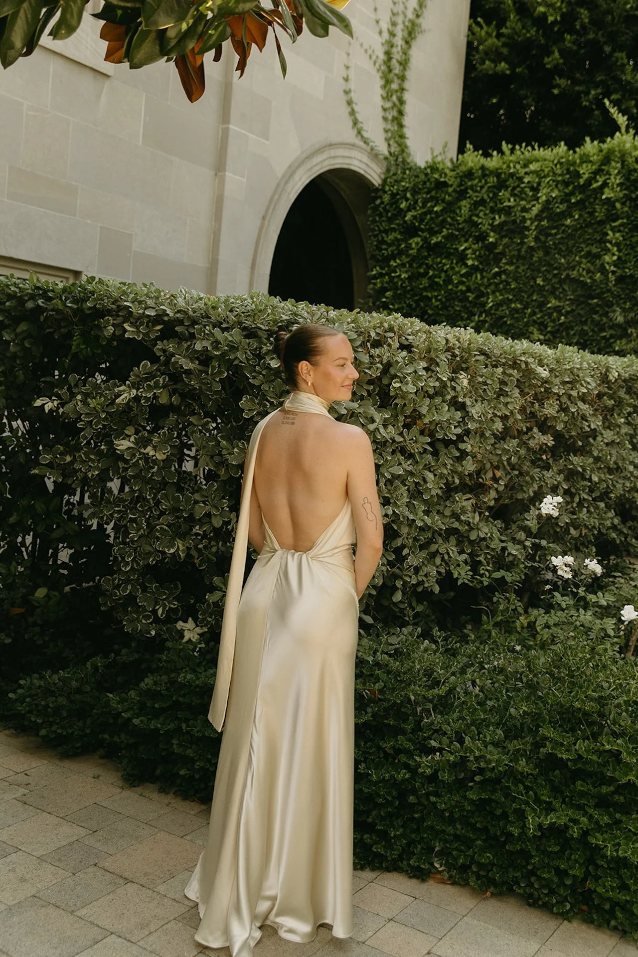 The bride standing in a garden courtyard with her back to the camera, showing the open-back satin gown and long scarf detail.