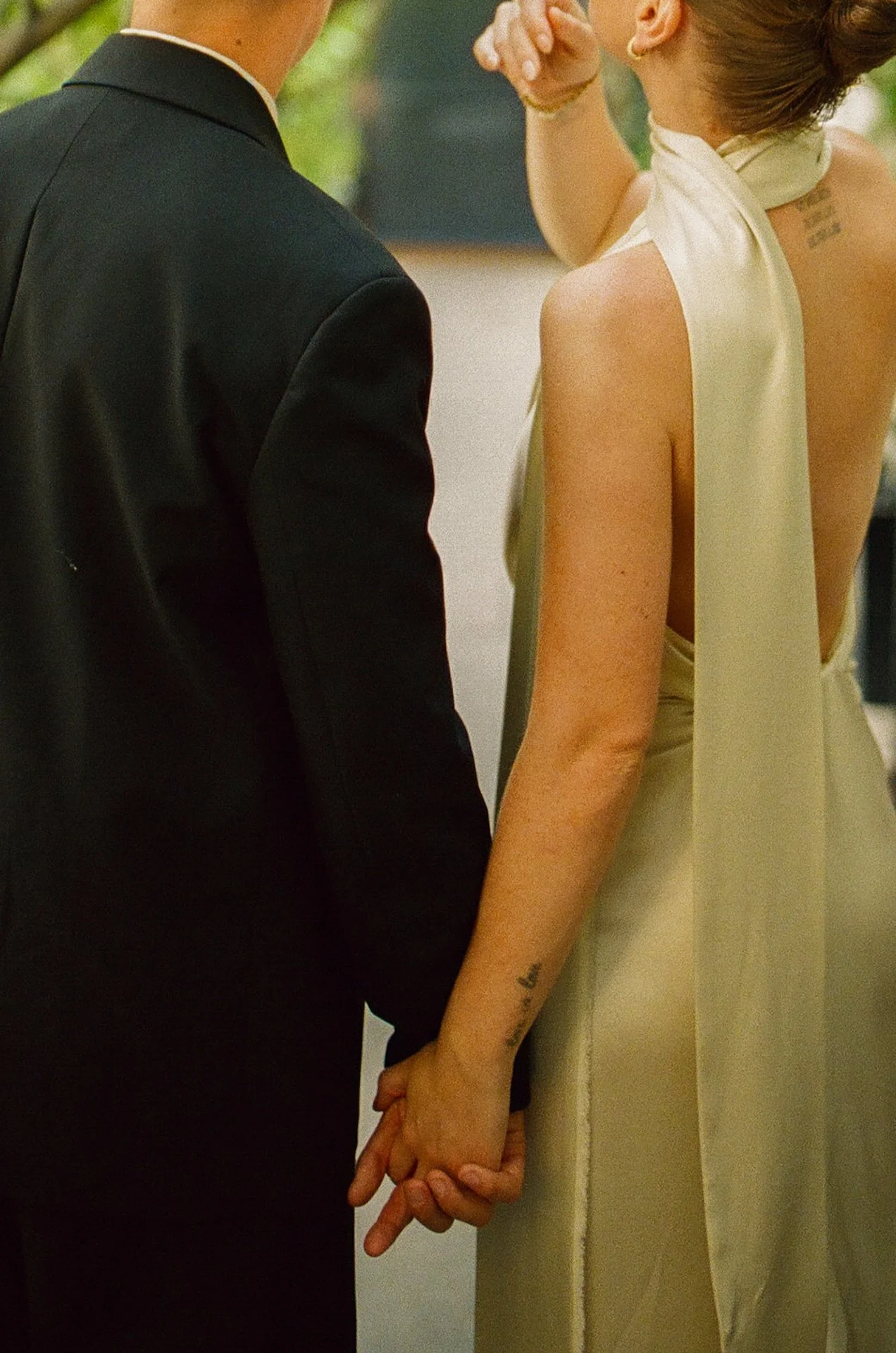 The couple holding hands while walking together outdoors, with the bride’s scarf detail flowing down her back and soft greenery surrounding them.