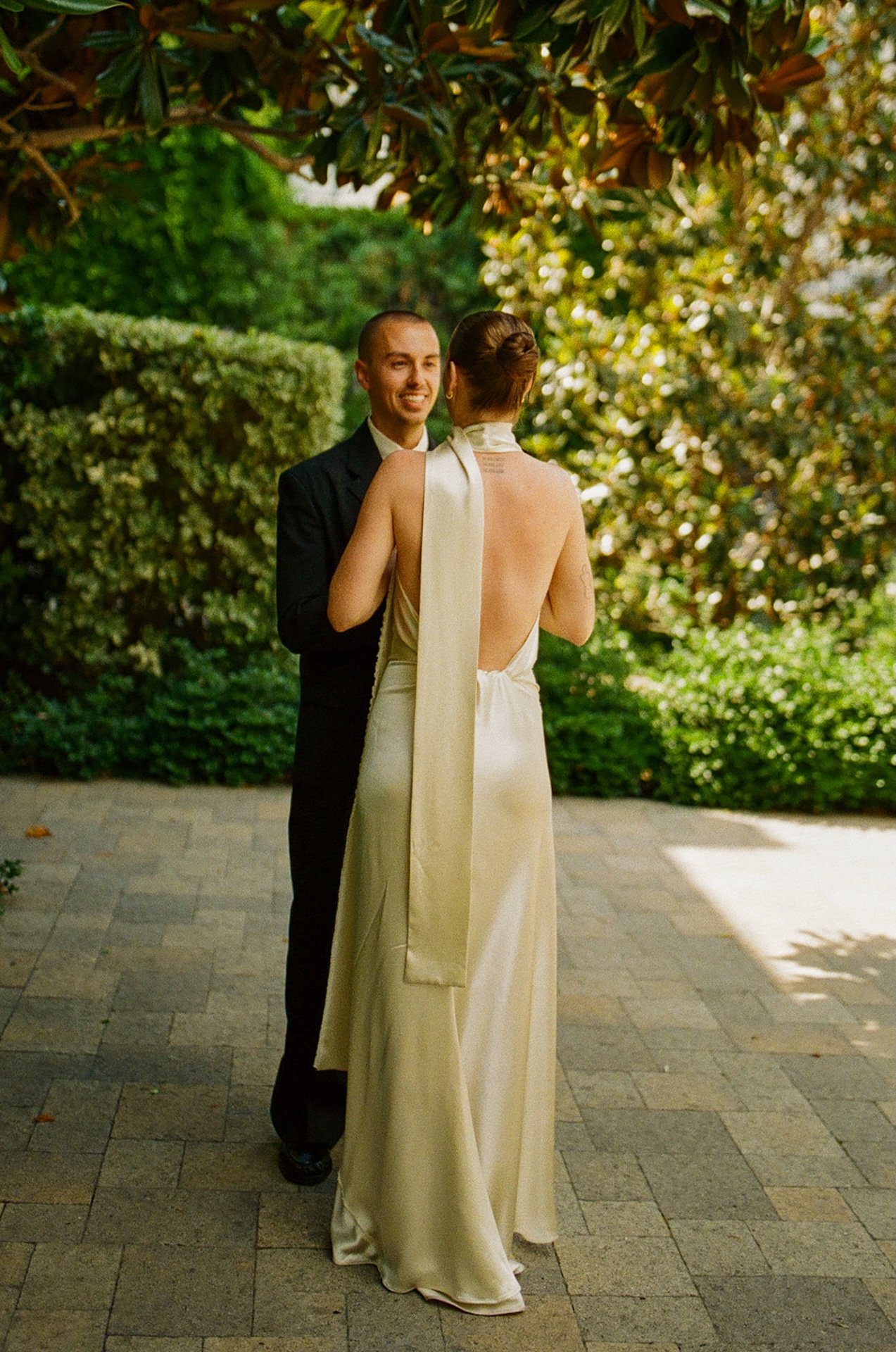 A full-body shot of the couple standing face to face in a garden courtyard, framed by greenery and soft natural light.