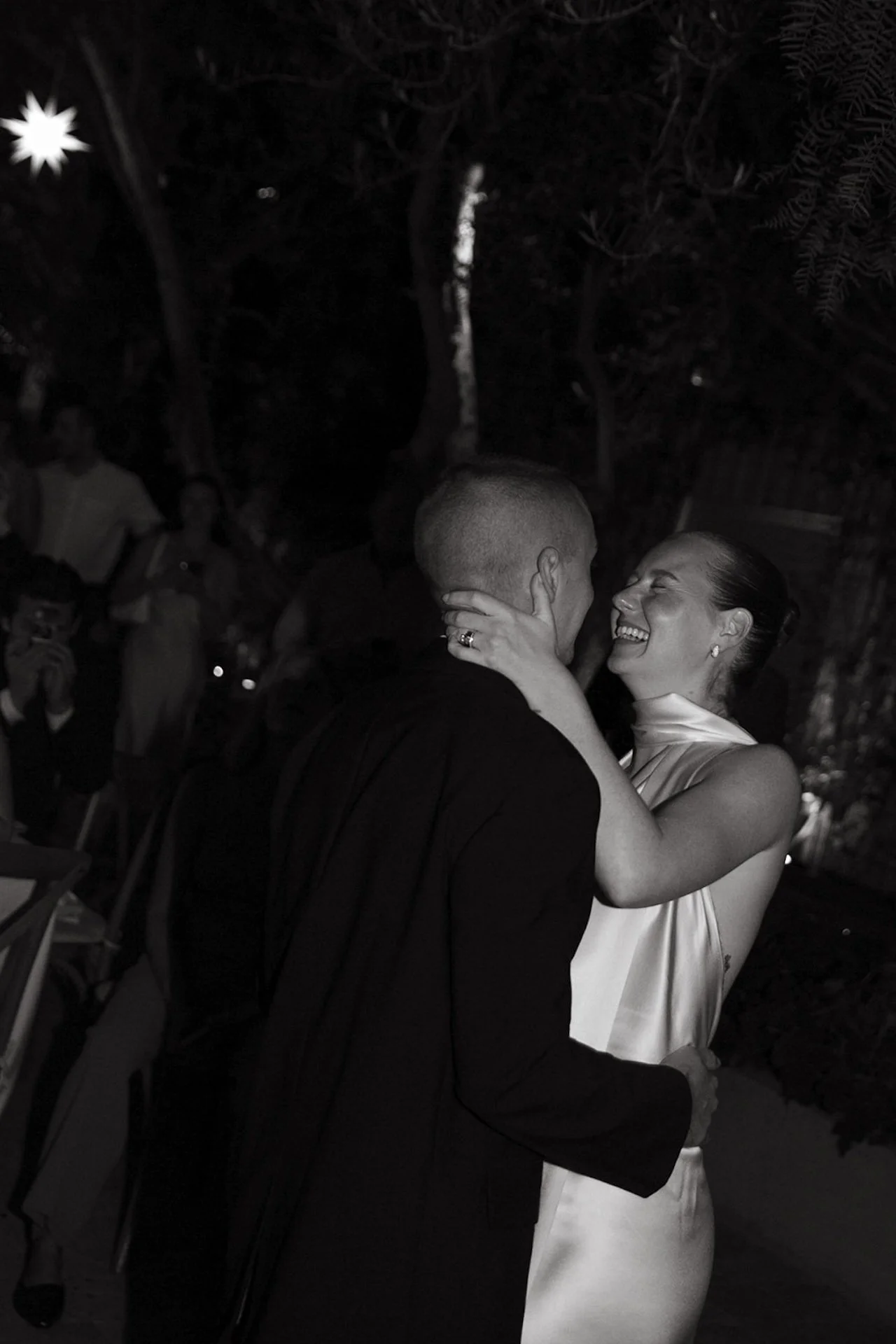 A black and white photo of a couple slow dancing during the reception, with the groom in a black suit and the bride in a satin halter gown, holding each other closely in a dimly lit outdoor setting.