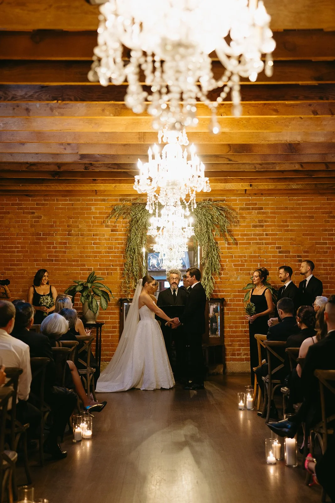 A symmetrical shot of a wedding ceremony at the altar, framed by glowing chandeliers and a lush greenery installation against a brick backdrop.