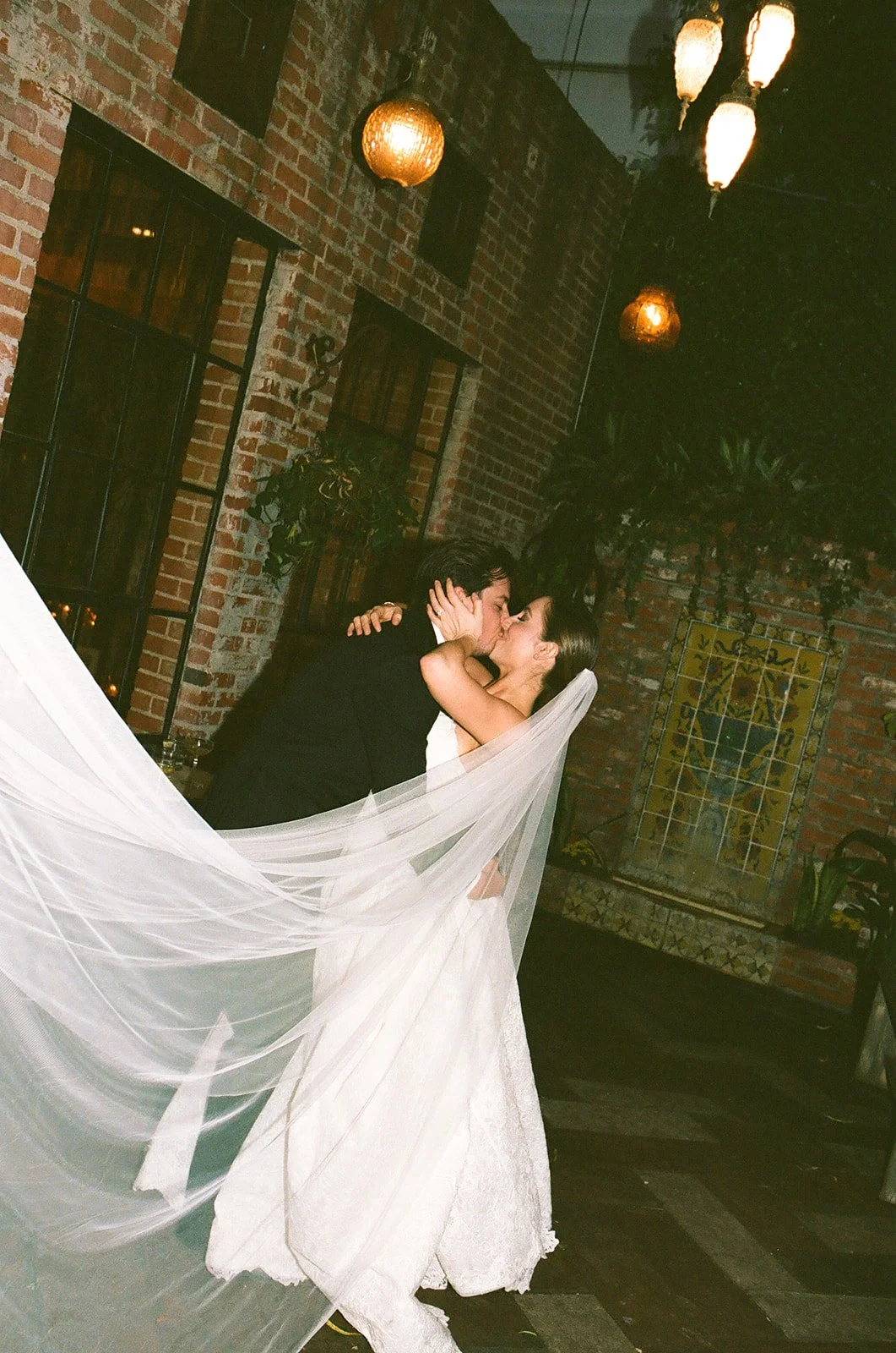 A film photo of a bride and groom kissing in a courtyard, with the bride's long white veil billowing in the foreground.