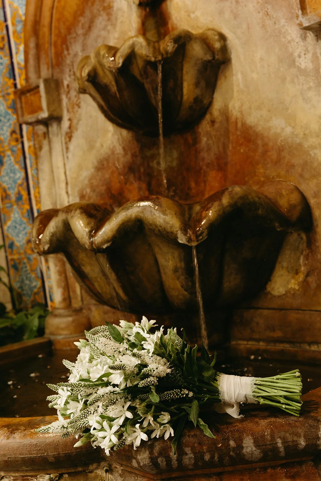 A bridal bouquet of white textured flowers and greenery resting on the edge of a vintage stone fountain at one of the top-rated DTLA wedding venues.
