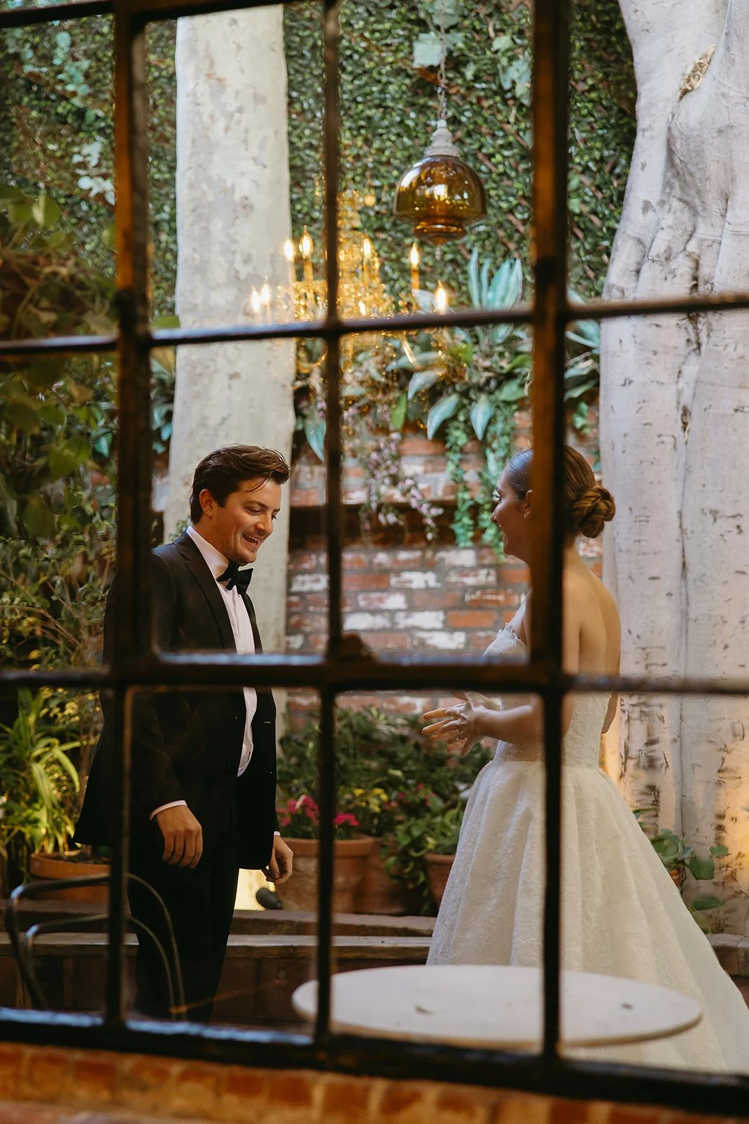 A groom looking at his bride with a smile, captured through a window pane with a lush, ivy-covered wall and a warm chandelier in the background.