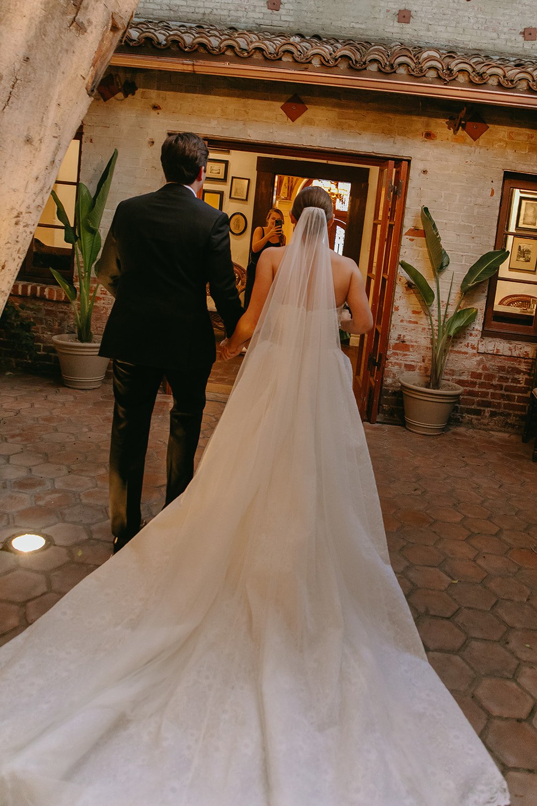 A rear view of a bride and groom walking hand-in-hand through a Mediterranean-style courtyard toward a set of open French doors.