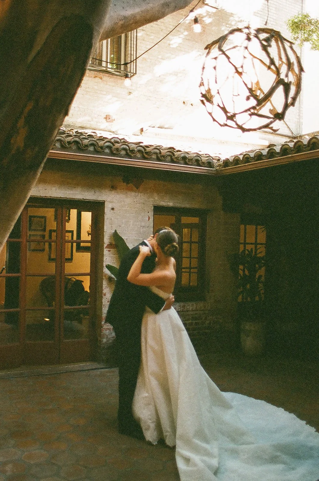 A bride and groom sharing a romantic embrace in a rustic outdoor courtyard at one of the premier DTLA wedding venues, featuring brick walls and a large statement tree.