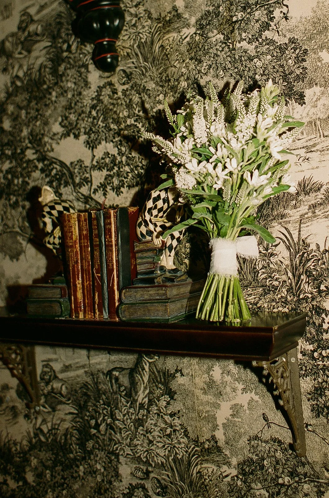 A detail photo of the brides white wedding florals sitting on a bookshelf with a vintage wallpaper behind it.