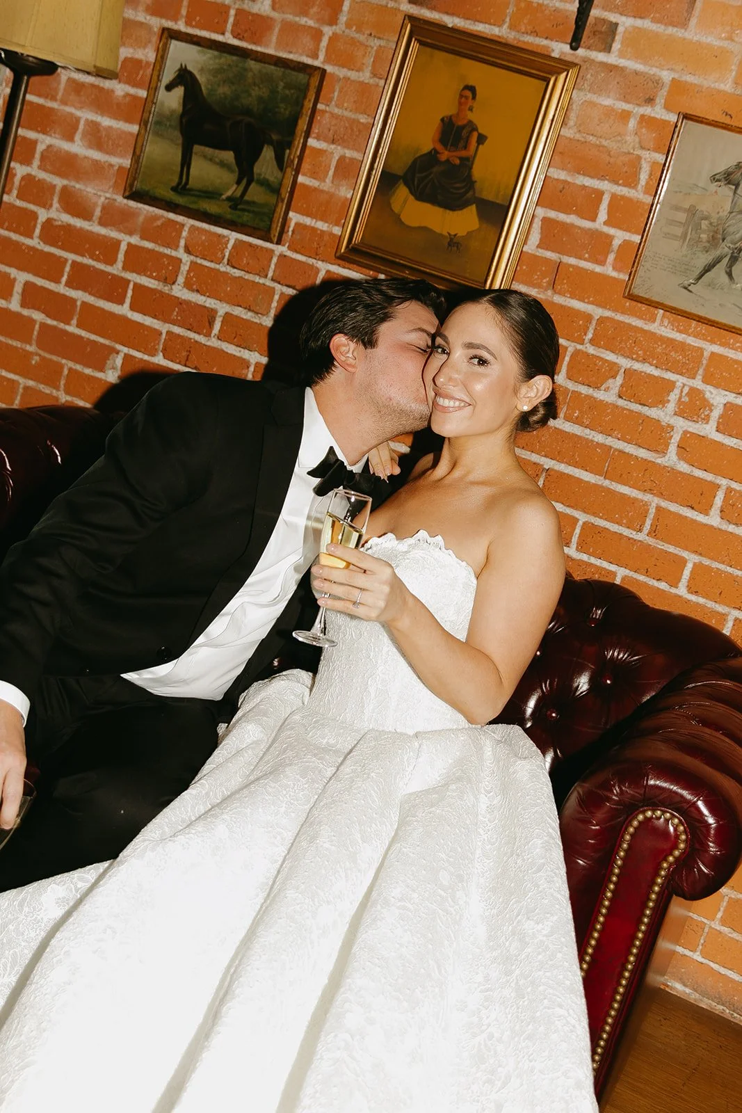 An elegant flash photo of the bride and groom sitting on a vintage couch together as the groom kisses his wife's cheek 