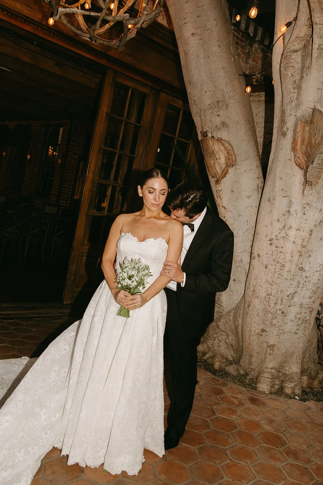 The bride and groom standing in the courtyard at their DTLA wedding venue as the bride stands holding her florals and the grom kisses her shoulder softly