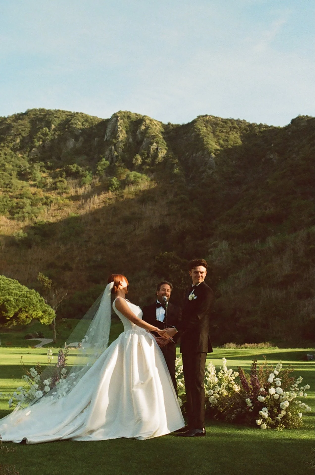 Bride and groom holding hands during their outdoor ceremony surrounded by florals and green canyon views in Laguna Beach.