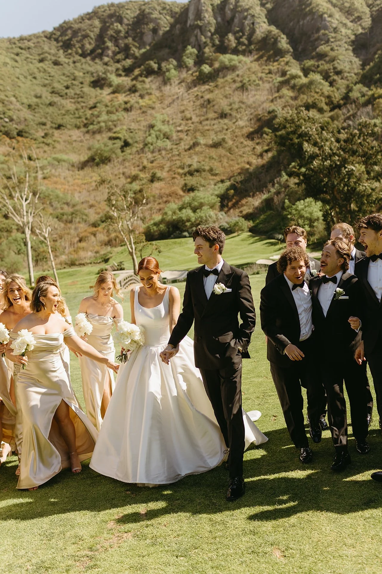 Wedding party walking together across the grass in champagne bridesmaid dresses and black tuxedos at The Ranch at Laguna Beach wedding.