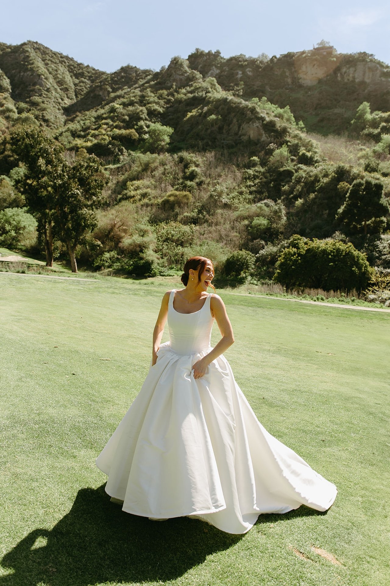 Bride twirling in her satin wedding dress on the lawn with canyon hills and sunshine behind her.