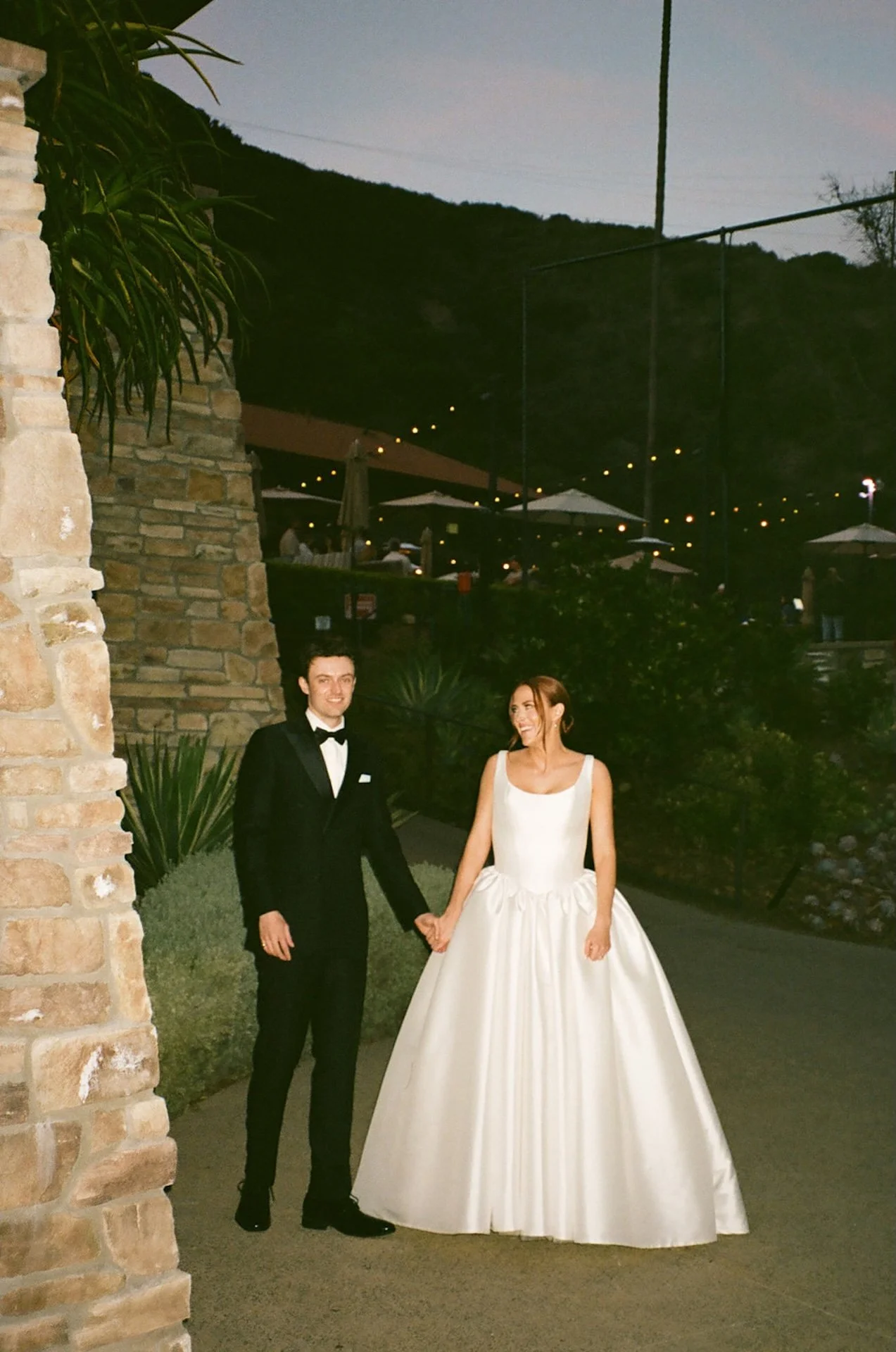 Bride and groom walking hand in hand through the venue at night surrounded by string lights and canyon views.