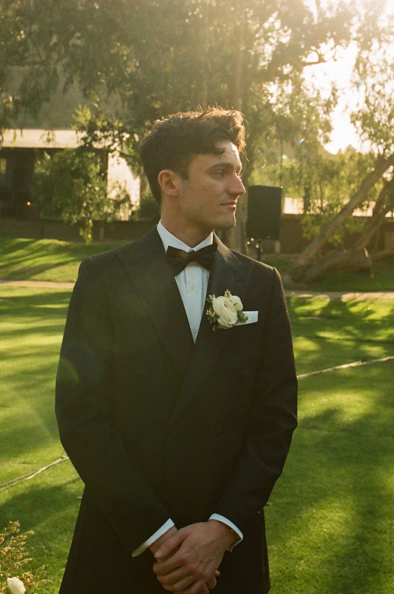 Groom portrait in a black tuxedo standing on the lawn with soft golden hour light and greenery behind him.