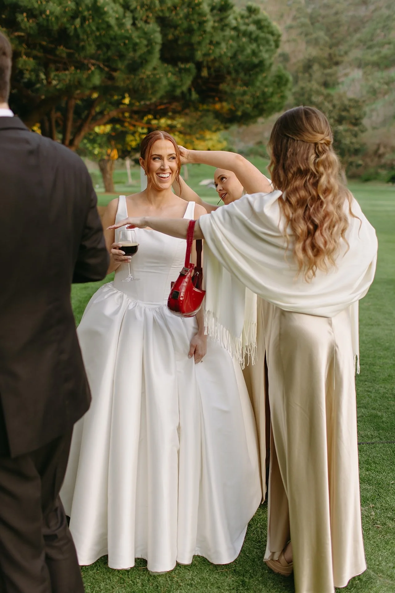 Bride chatting with guests in her satin wedding gown while a bridesmaid adjusts her hair during cocktail hour.