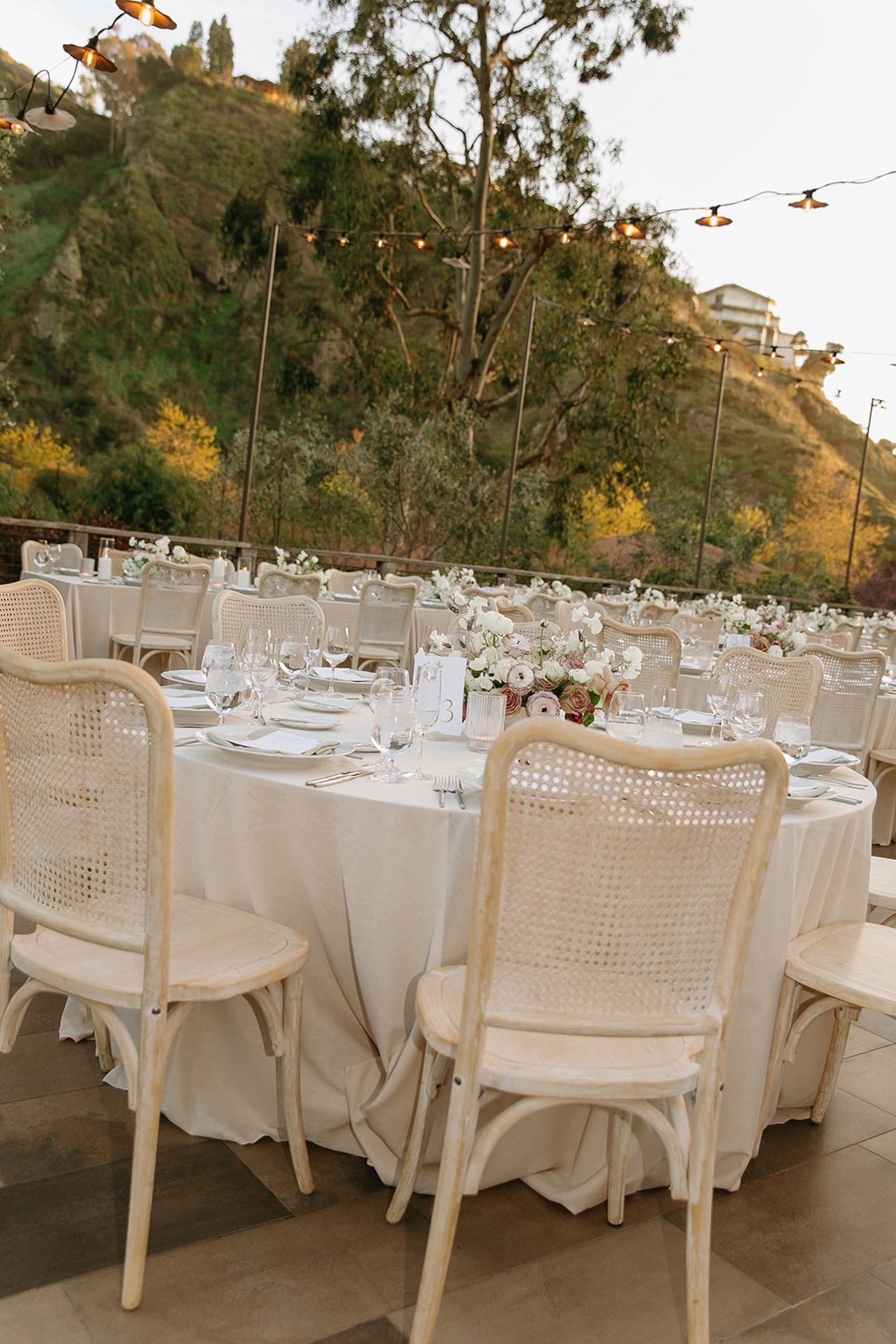 Outdoor reception tablescape with rattan chairs, neutral linens, and floral centerpieces beneath string lights.