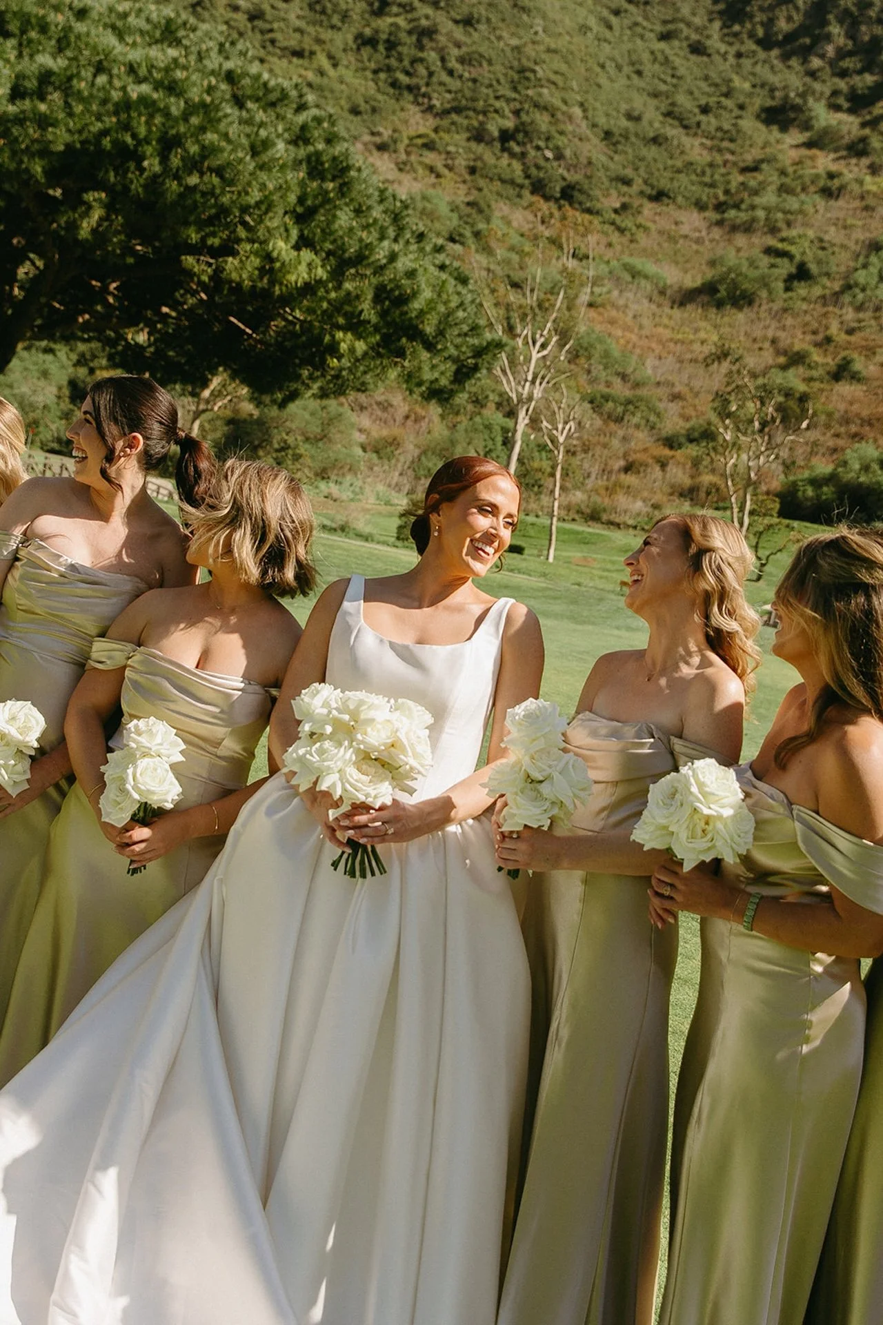 Bride laughing with bridesmaids in champagne dresses holding white rose bouquets outdoors.