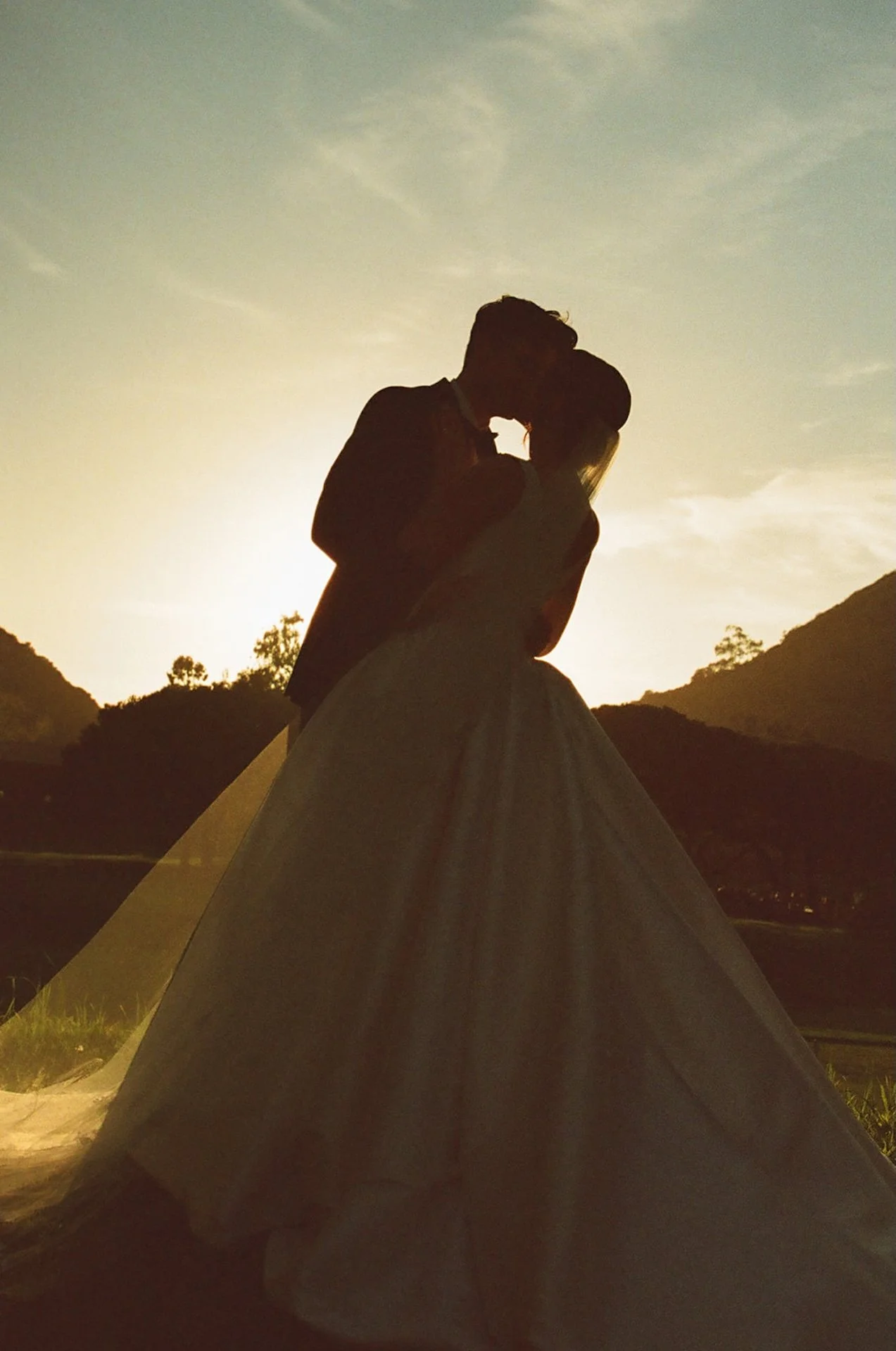 Silhouette portrait of bride and groom kissing against the sunset sky.