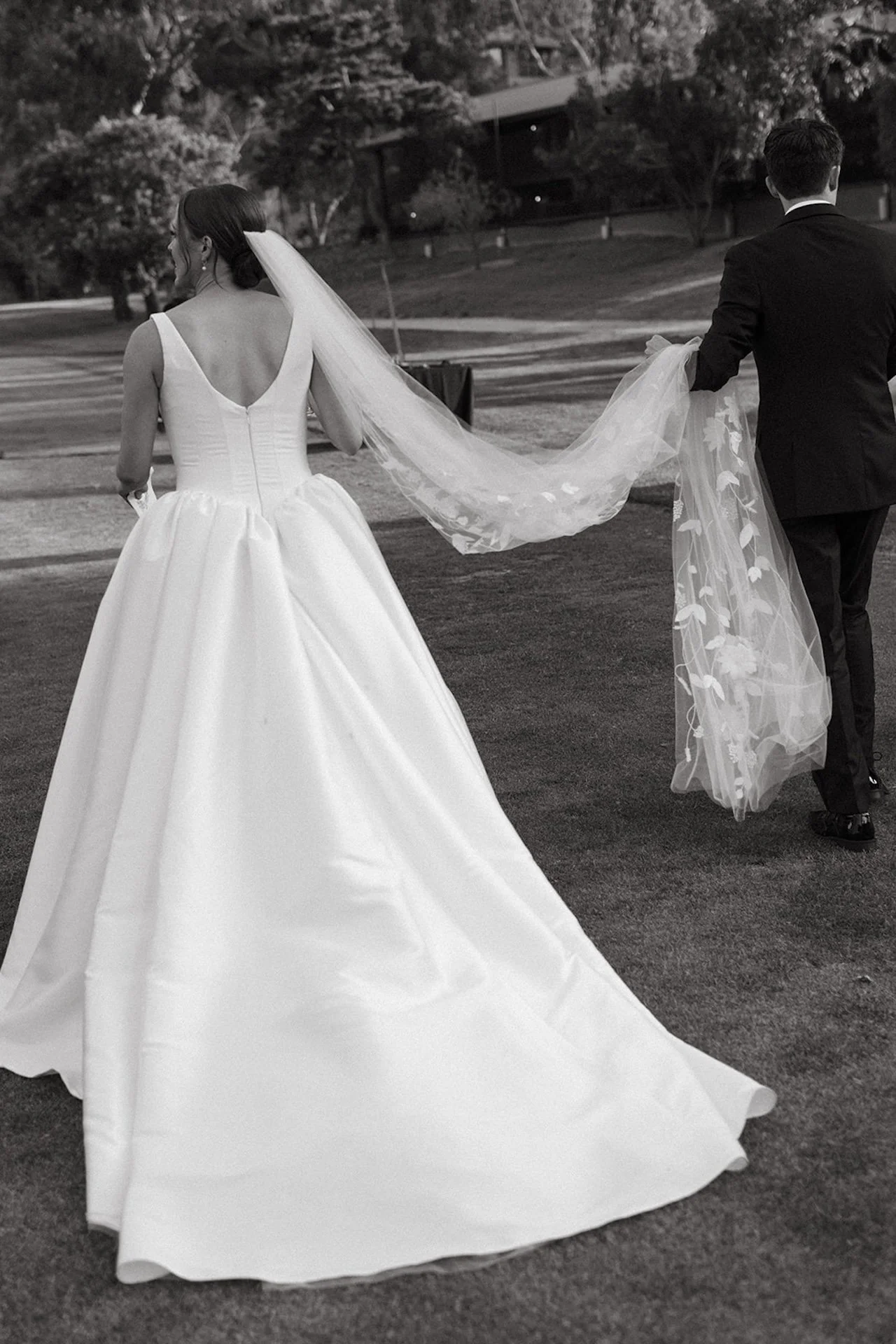 Black and white portrait of bride and groom walking across the lawn with her veil trailing behind at The Ranch at Laguna Beach wedding.