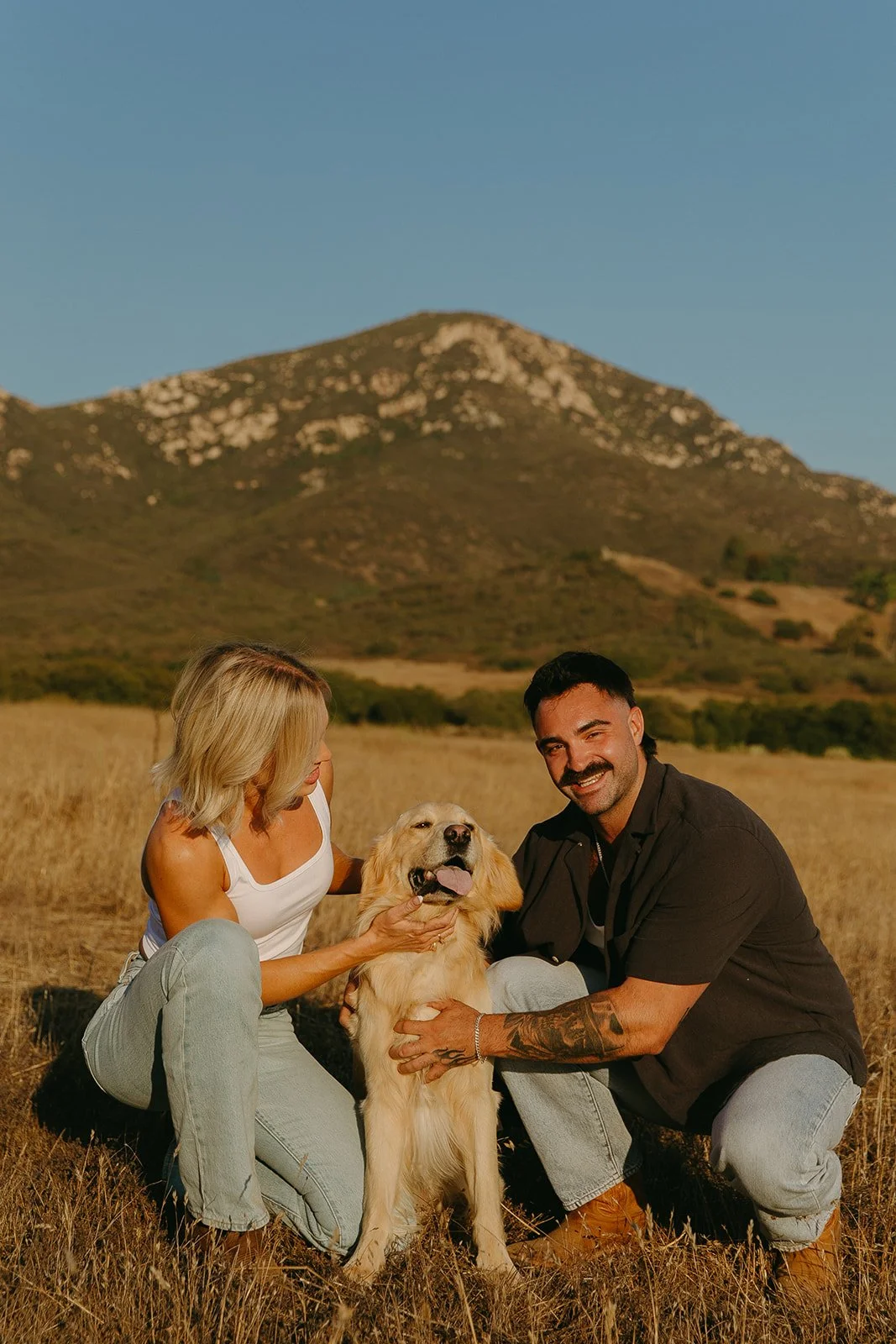 Couple kneeling in a sunlit field smiling with their golden retriever, mountain landscape behind them.