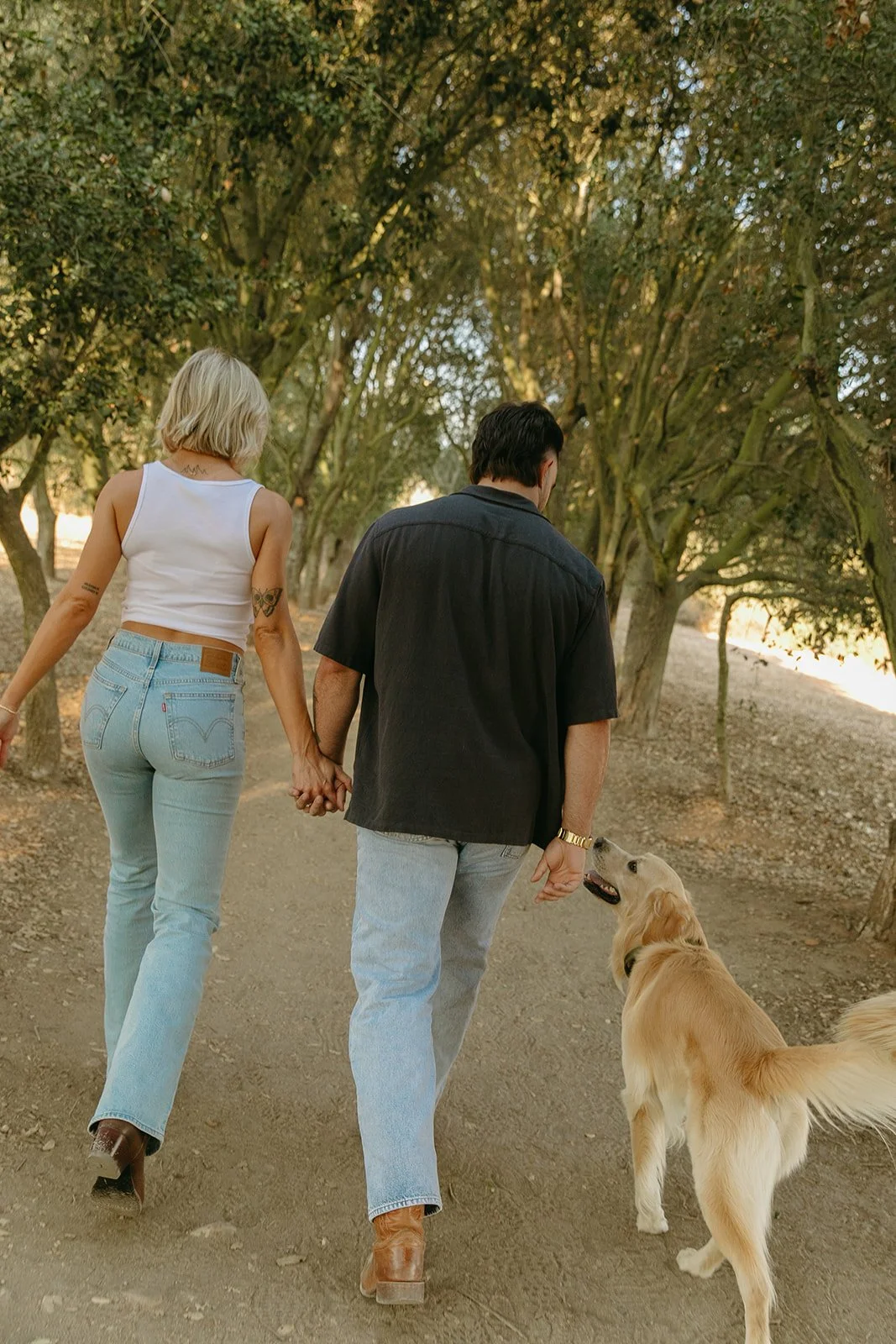 Walking western engagement photos from behind as the couple strolls through a shaded trail with their dog at their side.