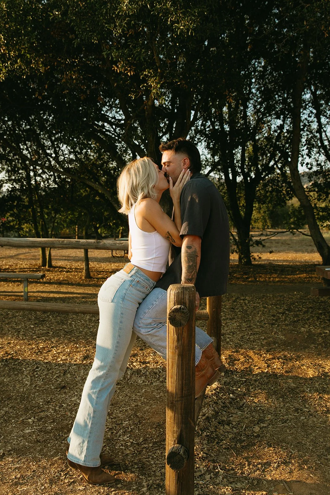 Golden hour western engagement photos of the couple kissing beside a rustic fence surrounded by oak trees.